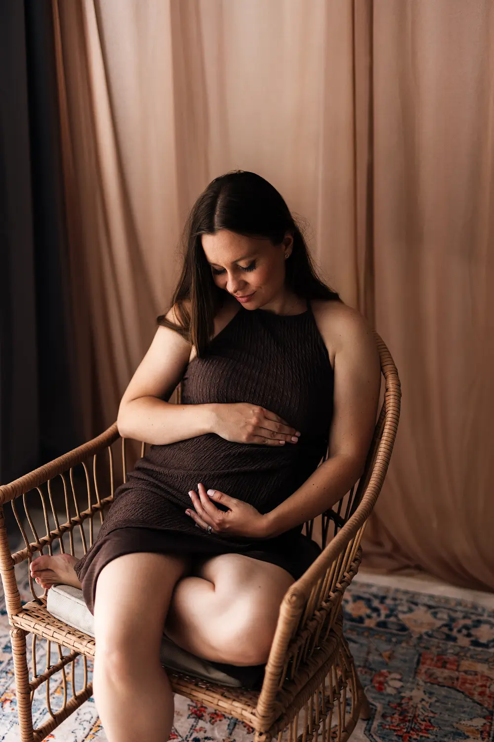 Tehotná žena si hladká bruško počas tehotenského fotenia v ateliéri pričom má oblečené hnede šaty na fotenie z klientskeho šatníka