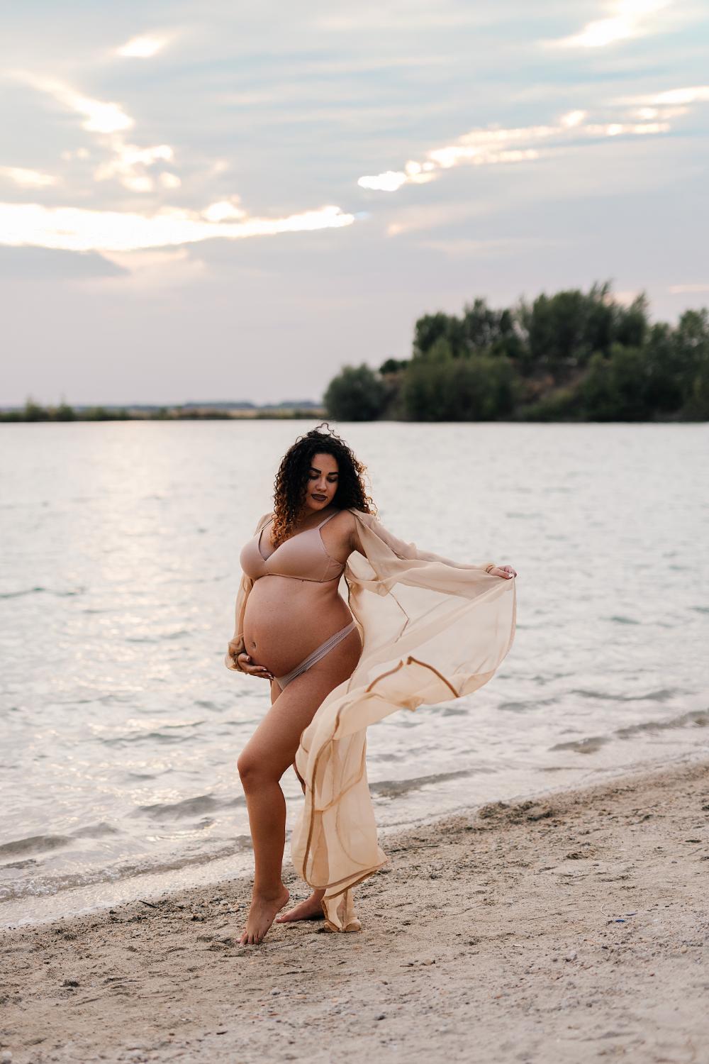 Tehotná žena stojí na pieskovej pláži pri jazere, jednou rukou drží tehotenské brucho, pričom druhou rukou drží jemný prehoz ktorý veje vo vetre pri západe slnka počas tehotenského fotenia v okolí Nitry 