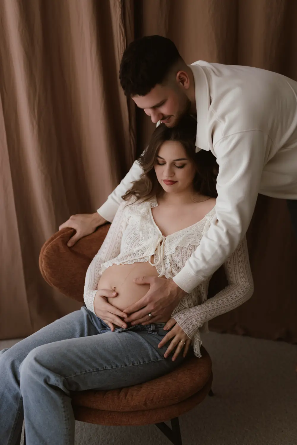  Muž drží tehotenské brucho ženy pričom tá sedí na stoličke počas uvoľneného tehotenského fotenia v ateliéri v Nitre 