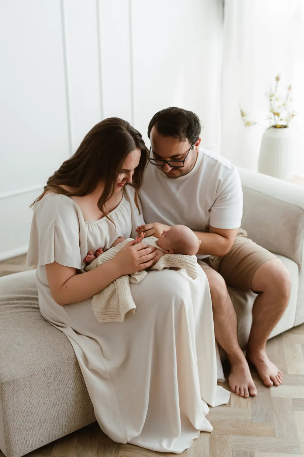 Rodičia sa usmievajú a hladkajú bábätko, pričom sedia na pohovke počas newborn fotenia v ateliéri v Šuranoch.