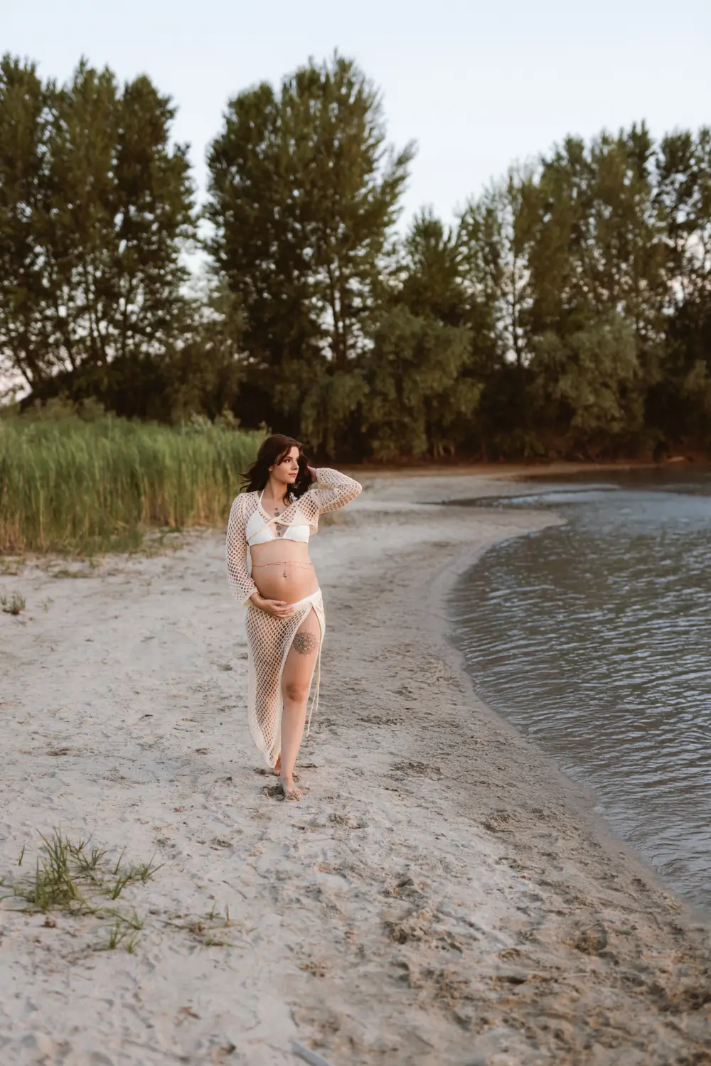 Tehotná žena sa prehádza po pláži pričom jemne pózuje počas tehotenského fotenia pri Západe slnka.